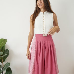 Puede incluir: Una blusa blanca sin mangas con cuello alto y botones dorados. La blusa se combina con una falda rosa con estampado floral. La falda tiene pliegues y una cintura blanca con una hebilla dorada.
