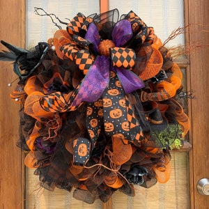 May include: A Halloween wreath with orange and black mesh, a purple and black checkered bow, and a black witch's hat. The wreath is decorated with orange and white jack-o'-lantern patterned ribbon.