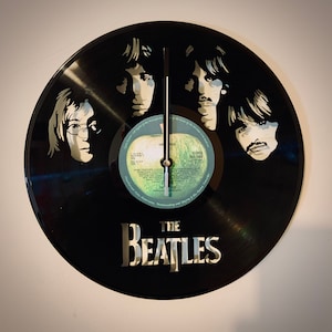 May include: A black vinyl record clock featuring the Beatles. The clock face has a green apple design with the text "Come Together" and "The Beatles" printed in white. The clock hands are black and the Beatles are depicted in black and white silhouettes.