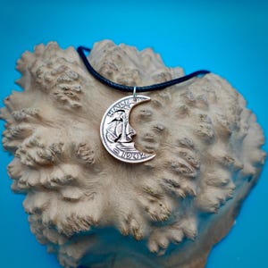 May include: A crescent moon pendant necklace made from a half British coin. The coin is dated 1974 and features a ship and the words "HALF CROWN". The necklace is on a black cord and is displayed on a piece of wood.