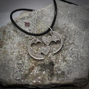 May include: Two silver heart-shaped pendants on a black cord necklace. The pendants are cut from old coins and are joined together to form a single heart.
