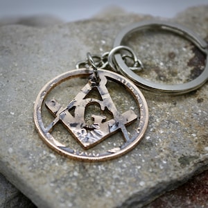 May include: A copper-colored key chain with a silver key ring. The key chain is a round coin with a square and compass symbol cut out of the center. The coin has the year 1960 engraved on it.