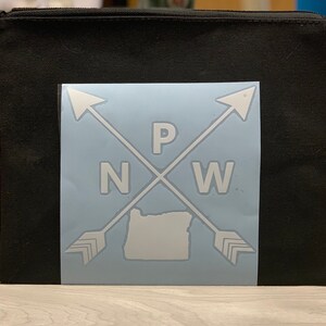 May include: A white vinyl sticker with a graphic of two arrows crossing over each other with the letters "P", "N", and "W" in the center. The state of Oregon is in the center of the graphic. The sticker is on a black canvas zippered pouch.