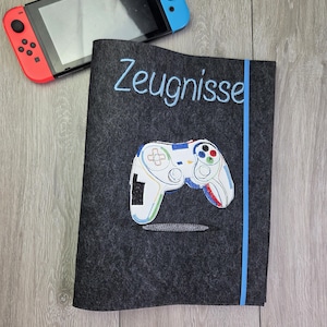 Könnte beinhalten: Eine dunkelgraue Filzhülle mit dem Wort "Zeugnisse" in Hellblau gestickt und einem farbenfrohen Videospiel-Controller-Design. Eine blau-rote Videospielkonsole befindet sich im Hintergrund.