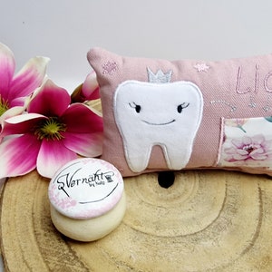 Könnte beinhalten: Ein rosa Zahnkissen mit einem aufgestickten weißen Zahn. Der Zahn hat eine Krone und ein lächelndes Gesicht. Das Kissen hat den Namen "Lia" aufgestickt. Das Kissen liegt auf einer Holzoberfläche mit einer kleinen Holzkiste mit rosa Deckel.