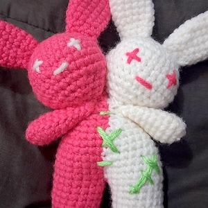 Puede incluir: Una muñeca de conejo de ganchillo hecha a mano, mitad rosa y mitad blanca. El lado rosa tiene ojos cosidos blancos, mientras que el lado blanco tiene ojos cosidos rosas. Las costuras verdes acentúan la costura. Un juguete de peluche único y artesanal.