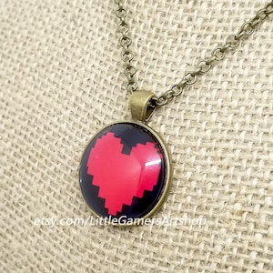 May include: A necklace with a bronze chain and a round pendant featuring a pixelated red heart on a black background. The pendant is hanging on a burlap fabric background.