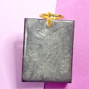 May include: A silver glitter resin keychain with a gold key ring. The keychain is rectangular and has a smooth, shiny surface.
