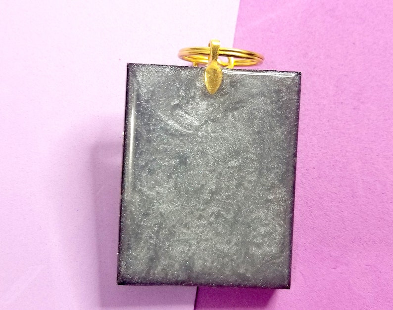 May include: A silver glitter resin keychain with a gold key ring. The keychain is rectangular and has a smooth, shiny surface.
