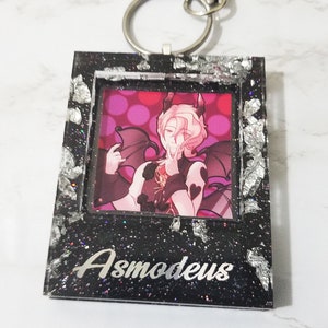 May include: Black and silver glitter frame keychain with a picture of a pink-haired anime character with wings and the text "Asmodeus" on the bottom.