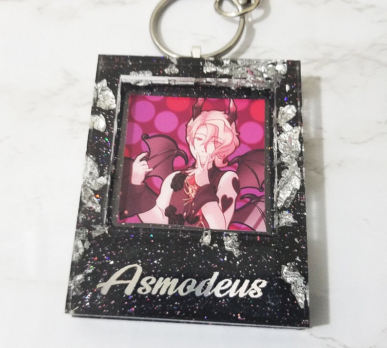 May include: Black and silver glitter frame keychain with a picture of a pink-haired anime character with wings and the text "Asmodeus" on the bottom.