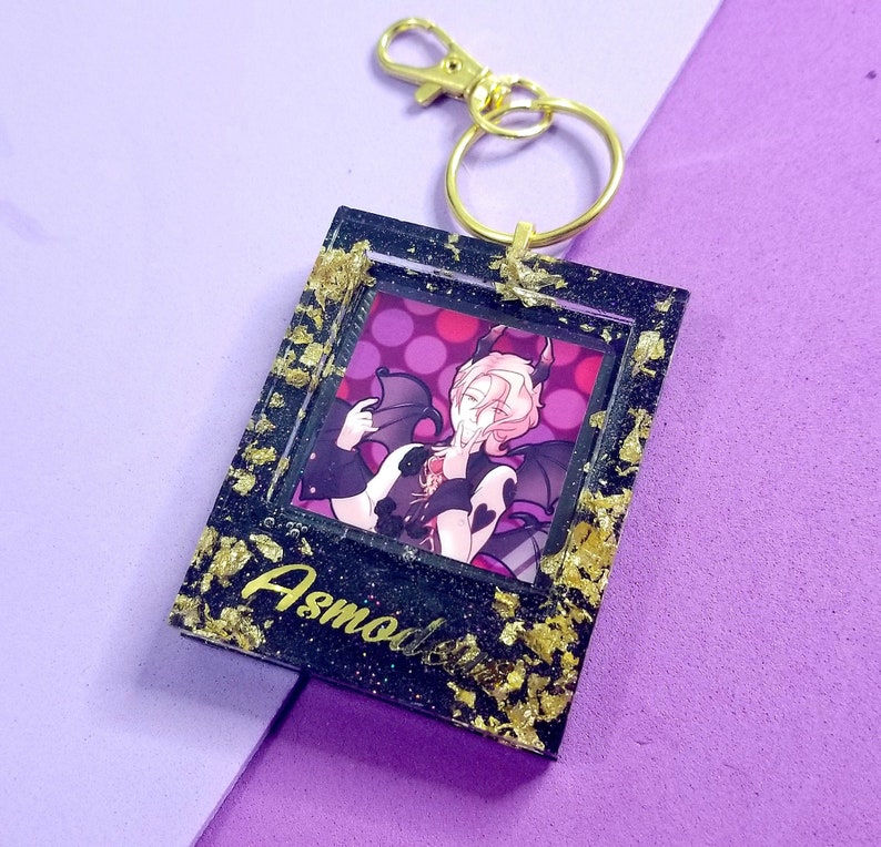 May include: A black and gold keychain with a picture of a character with pink hair and wings. The character is holding a heart. The keychain has the word "Asmodeus" written on it.