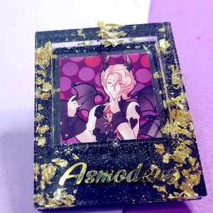 May include: A black square resin frame with gold glitter and gold leaf accents. The frame contains a printed image of a character with pink hair and devil horns. The text "Asmodeus" is written in gold lettering at the bottom of the frame.