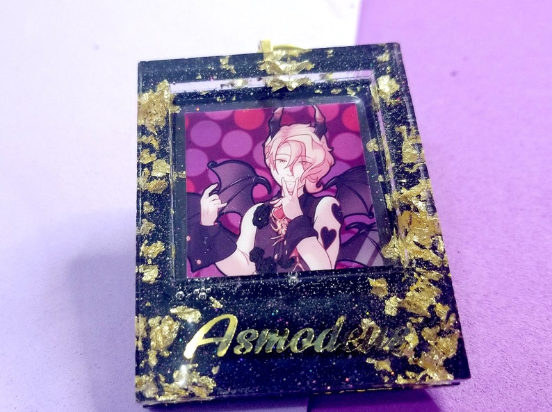 May include: A black square resin frame with gold glitter and gold leaf accents. The frame contains a printed image of a character with pink hair and devil horns. The text "Asmodeus" is written in gold lettering at the bottom of the frame.