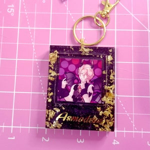 May include: A black and gold resin keychain featuring a character with pink hair and wings. The character is wearing a black and white outfit and has a pink background with white polka dots. The keychain has the text "Asmodeus" in gold lettering.