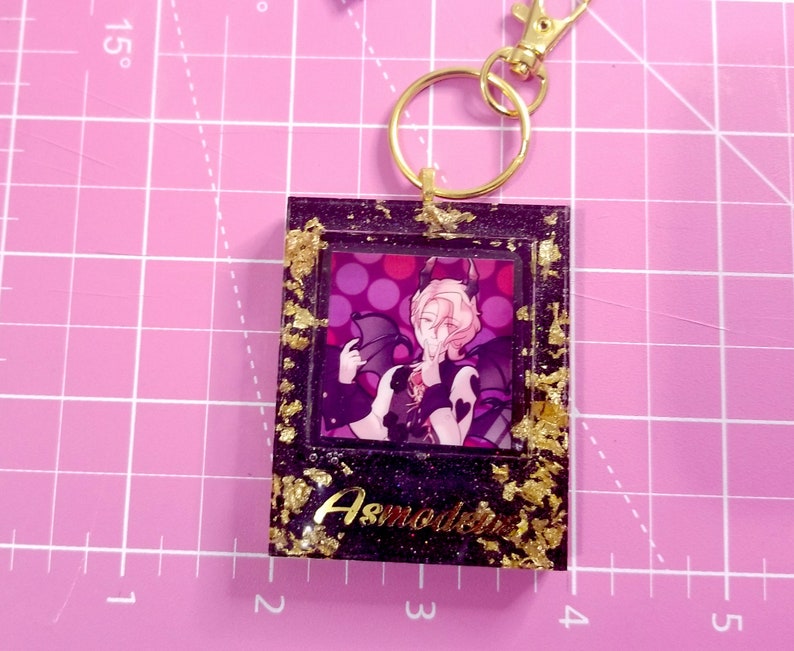May include: A black and gold resin keychain featuring a character with pink hair and wings. The character is wearing a black and white outfit and has a pink background with white polka dots. The keychain has the text "Asmodeus" in gold lettering.