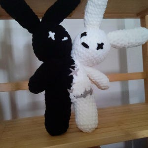 May include: Two crocheted bunny plushies, one black and one white, are sewn together to create a two-toned bunny. The black bunny has a stitched-on X for eyes and the white bunny has a stitched-on X for eyes and a stitched-on mouth.