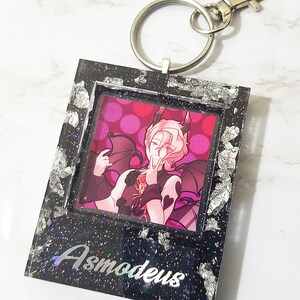 May include: Black and silver glitter resin keychain with a picture of a character with pink hair and wings. The text "Asmodeus" is written on the bottom of the keychain.