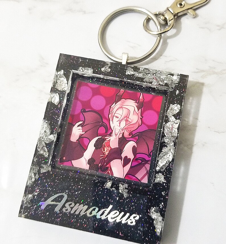 May include: Black and silver glitter resin keychain with a picture of a character with pink hair and wings. The text "Asmodeus" is written on the bottom of the keychain.