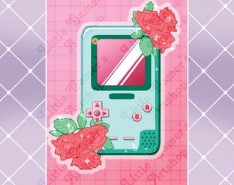 May include: A teal and green handheld video game console with a pink screen and buttons. The console is surrounded by pink roses with green leaves and glitter.