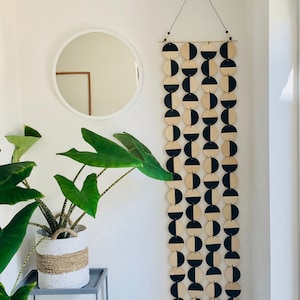 Puede incluir: Un tapiz de pared geométrico de madera negra y natural con un patrón de círculos y semicírculos. El tapiz está hecho de madera y está suspendido del techo con una cuerda.