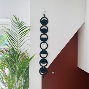 Op de afbeelding: Een zwarte metalen maanfase wanddecoratie met acht verschillende maanfasen. De decoratie is bevestigd aan een witte muur met een klein haakje.