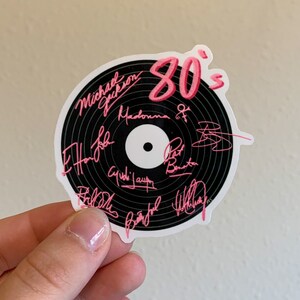 May include: A black vinyl record sticker with pink text that says "80's" and includes the names of popular artists from the 1980s, including Michael Jackson, Madonna, and Phil Collins.