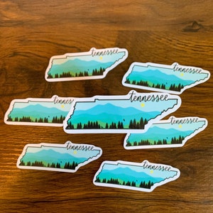 May include: Seven white stickers with a blue and green outline of the state of Tennessee. Each sticker has the word "Tennessee" written in black cursive script.