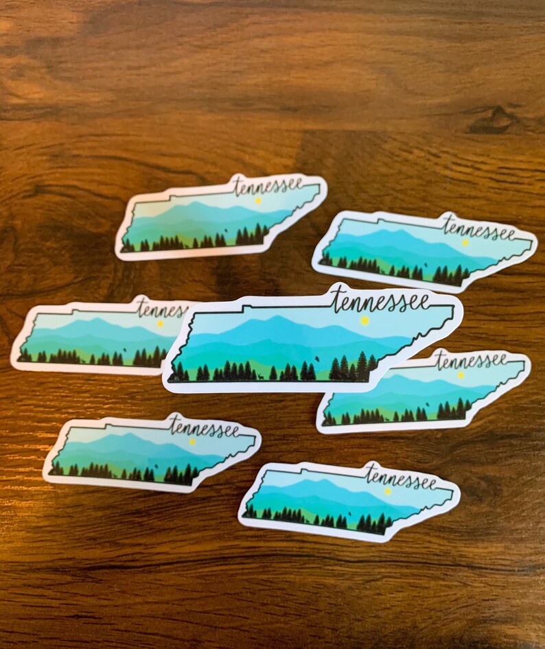 May include: Seven white stickers with a blue and green outline of the state of Tennessee. Each sticker has the word "Tennessee" written in black cursive script.