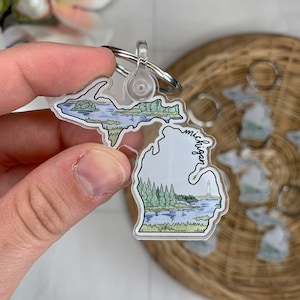 Puede incluir: Un llavero de acrílico transparente con forma del estado de Michigan. El llavero presenta una ilustración de acuarela de la península superior y la península inferior con un faro en la península inferior. La palabra "Michigan" está escrita verticalmente en el lateral del llavero.