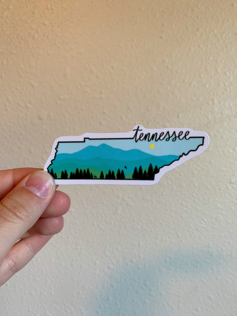 May include: A white sticker with a blue and green outline of the state of Tennessee. The sticker features a mountain range with a small yellow sun and the word "Tennessee" written in black cursive.