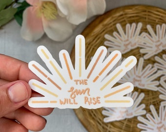 May include: A white sticker with a sunburst design in shades of orange and yellow. The text "THE sun WILL RISE" is written in a handwritten font.