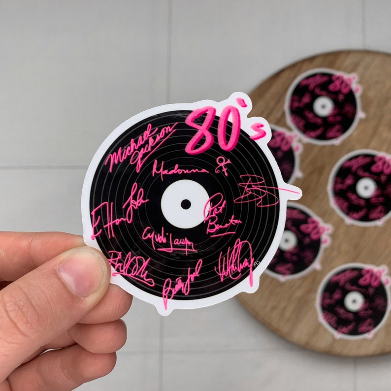May include: A black and white vinyl record sticker with pink 80's text. The sticker features the signatures of popular 80's musicians including Michael Jackson, Madonna, and Phil Collins.