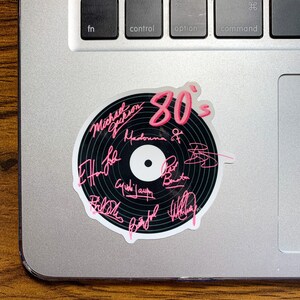 May include: A black vinyl record sticker with the text "80's" in pink lettering. The sticker features the signatures of various 80's music artists, including Michael Jackson, Madonna, and Phil Collins.