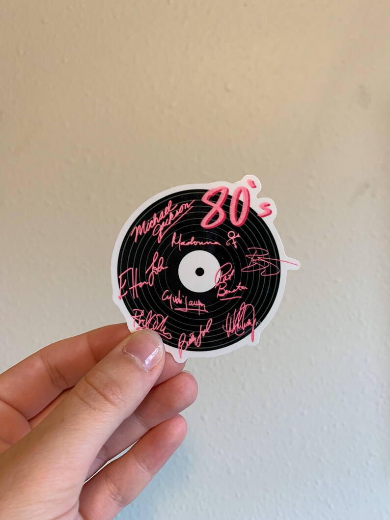 May include: A black vinyl record sticker with pink text that says "80's" and includes the names of popular artists from the 1980s, including Michael Jackson, Madonna, and Phil Collins.