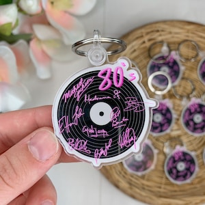 May include: A clear acrylic keychain shaped like a vinyl record with the text "80's" in pink lettering. The record is black with a white center and features pink handwritten signatures of popular 80's musicians around the edge.