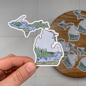 May include: A sticker of the state of Michigan with a lighthouse and trees on the lower peninsula and a lake scene on the upper peninsula. The sticker is white with a blue and green watercolor design. The word "Michigan" is written on the side of the lower peninsula.