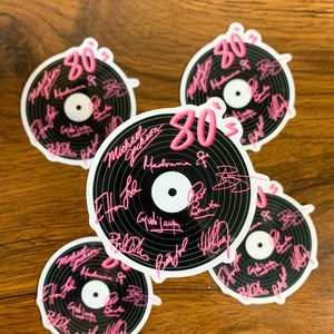 May include: Five black and white vinyl record stickers with pink 80's text. The stickers feature the signatures of popular 80's musicians including Michael Jackson, Madonna, and Billy Joel.
