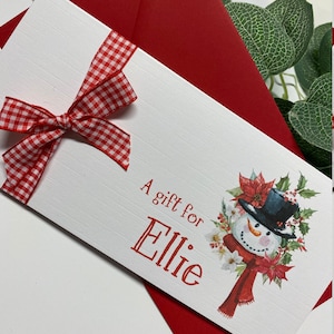 May include: A white gift card with a red and white gingham bow. The card has a snowman illustration with a red scarf and a wreath of holly and poinsettias. The text "A gift for Ellie" is printed on the card.