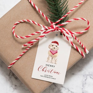 Pode incluir: Uma etiqueta de presente branca com uma ilustração em aquarela de um cão usando um chapéu de Papai Noel e uma bandana vermelha. O texto na etiqueta diz "Feliz Natal" em letras vermelhas cursivas e "Com amor da família Stephens" em letras pretas.