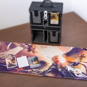 Può includere: Un organizzatore di giochi di carte nero con maniglia e più scomparti. L'organizzatore è seduto su un tavolo con un grande tappetino da gioco che presenta un'illustrazione fantasy di un guerriero alato con una luce brillante alle sue spalle. Il tappetino ha uno schema di colori viola, rosa e arancione.