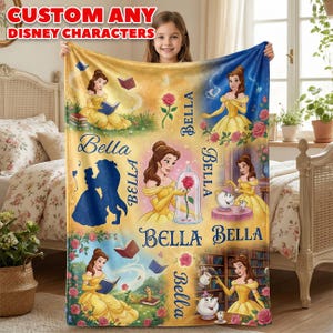 May include: A soft fleece blanket featuring multiple illustrations of the character Belle from Beauty and the Beast, including scenes with a rose, books, and the Beast. The text 'BELLA' is printed repeatedly across the yellow and blue background.