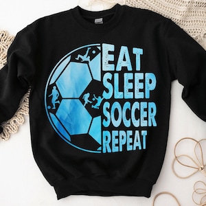 K&ouml;nnte beinhalten: Schwarzes Sweatshirt mit einem blauen und wei&szlig;en Fu&szlig;ball-Motiv und dem Text "Eat Sleep Soccer Repeat".