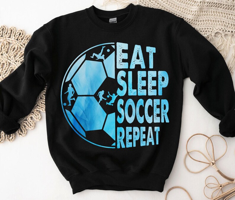 K&ouml;nnte beinhalten: Schwarzes Sweatshirt mit einem blauen und wei&szlig;en Fu&szlig;ball-Motiv und dem Text "Eat Sleep Soccer Repeat".