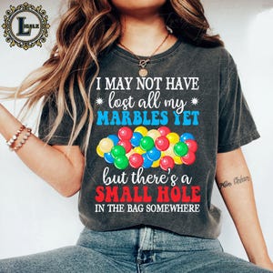 May include: Dark gray t-shirt with the text "I MAY NOT HAVE lost all my MARBLES YET but there's a SMALL HOLE IN THE BAG SOMEWHERE." A colorful graphic of marbles is below the text. The shirt is a casual style.