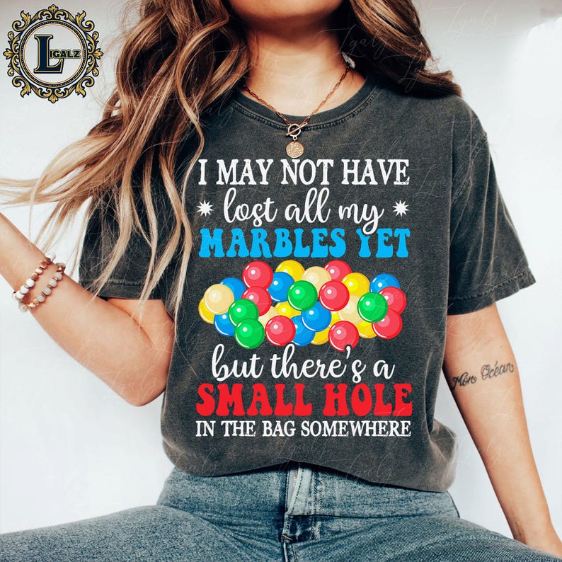 May include: Dark gray t-shirt with the text "I MAY NOT HAVE lost all my MARBLES YET but there's a SMALL HOLE IN THE BAG SOMEWHERE." A colorful graphic of marbles is below the text. The shirt is a casual style.