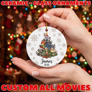 May include: A round white ceramic ornament featuring a Zootopia character graphic, including Judy Hopps, Nick Wilde, and others in a tree formation. The ornament displays the name James and the year 2026, accented with gold snowflakes.