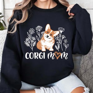 以下が含まれることがあります： 花柄と「Corgi Mom」の文字、ハートマークがプリントされた白いグラフィックが施された黒いスウェットシャツ。グラフィックには、コーギー犬が描かれています。
