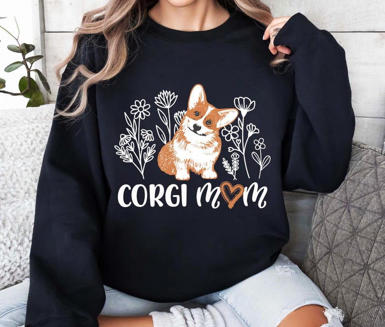 以下が含まれることがあります： 花柄と「Corgi Mom」の文字、ハートマークがプリントされた白いグラフィックが施された黒いスウェットシャツ。グラフィックには、コーギー犬が描かれています。