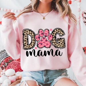 以下が含まれることがあります： 「Dog Mama」と書かれた、レオパード柄と肉球のプリントが施されたピンク色のスウェットシャツです。
